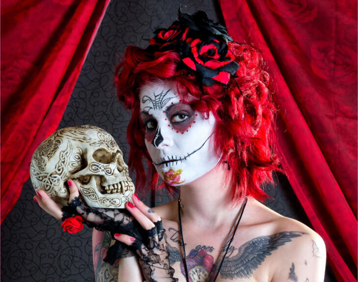 Frau mit Dia-de-los-Muertos-Gesichtsmaske und rotem Haar, hält einen verzierten Schädel in der Hand. Die Totenkopf-Frau (La Catrina) gilt als beliebtes Kostüm-Motiv. Das Gesichts-Make-Up ist inspiriert von den Zuckerschädeln (Calvera de Dulche) © Dirk Meeves