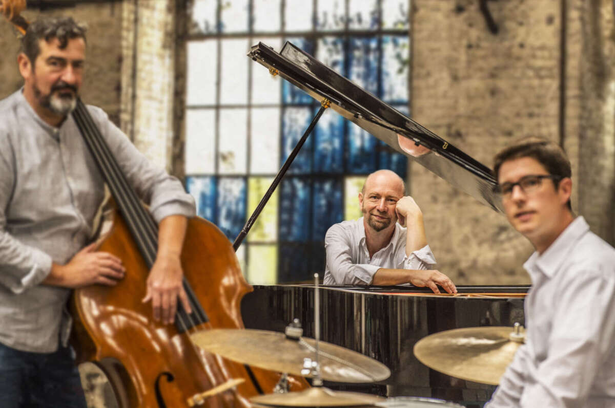 Edgar Knecht Trio mit Flügel, Kontrabass und Schlagzeug in einem hellen Raum mit großen Fenstern.
