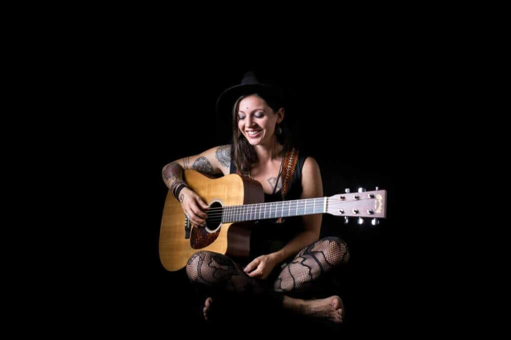 Singer-Songwriterin Nadine Fingerhut sitzt mit Akustikgitarre vor schwarzem Hintergrund und lächelt während eines Porträtshootings.| (c) Nadine Fingerhut