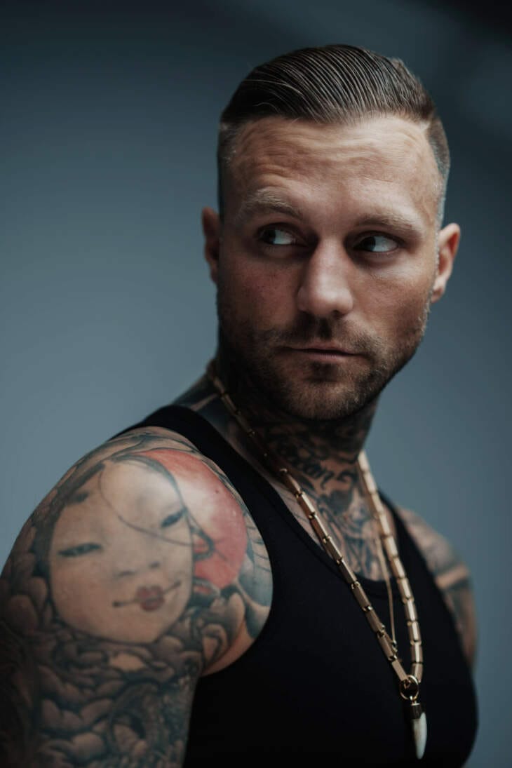 Nahaufnahme von Kontra K, dem Berliner Rapper, mit eindrucksvollen Tattoos und einem intensiven Blick zur Seite, er trägt ein schwarzes Tanktop und eine auffällige Kette, seine Tattoos auf Schulter und Hals sind prominent zu sehen Kontra K ist aktuell auf Tour. Hier trägt ein schwarzes Tanktop und eine markante Kette. Die intensiven Gesichtszüge und der fokussierte Blick betonen seine Persönlichkeit und die Tiefe seiner Musik. | (c) Niculai Constantinescu