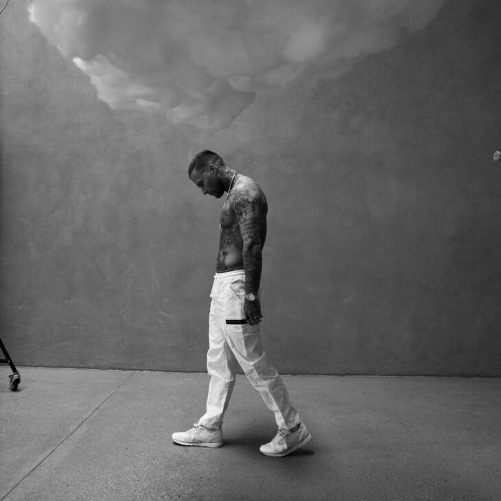 Kontra K in einer nachdenklichen Pose – der Berliner Rapper zeigt sich nachdenklich und verletzlich. Im Bild trägt er weiße Hosen und Sneaker, die Tattoos auf seinem Oberkörper sind gut sichtbar, während er mit gesenktem Kopf durch eine leere Szenerie geht Kontra K, der tätowierter Rapper und Musiker, steht nachdenklich und leicht nach vorne gebeugt in einem minimalistischen Setting. Das Bild zeigt ihn in einer intensiven und nachdenklichen Pose, was seine persönliche und künstlerische Tiefe betont. | Pressebild 03 - 2024 | (c) Niculai Constantinescu