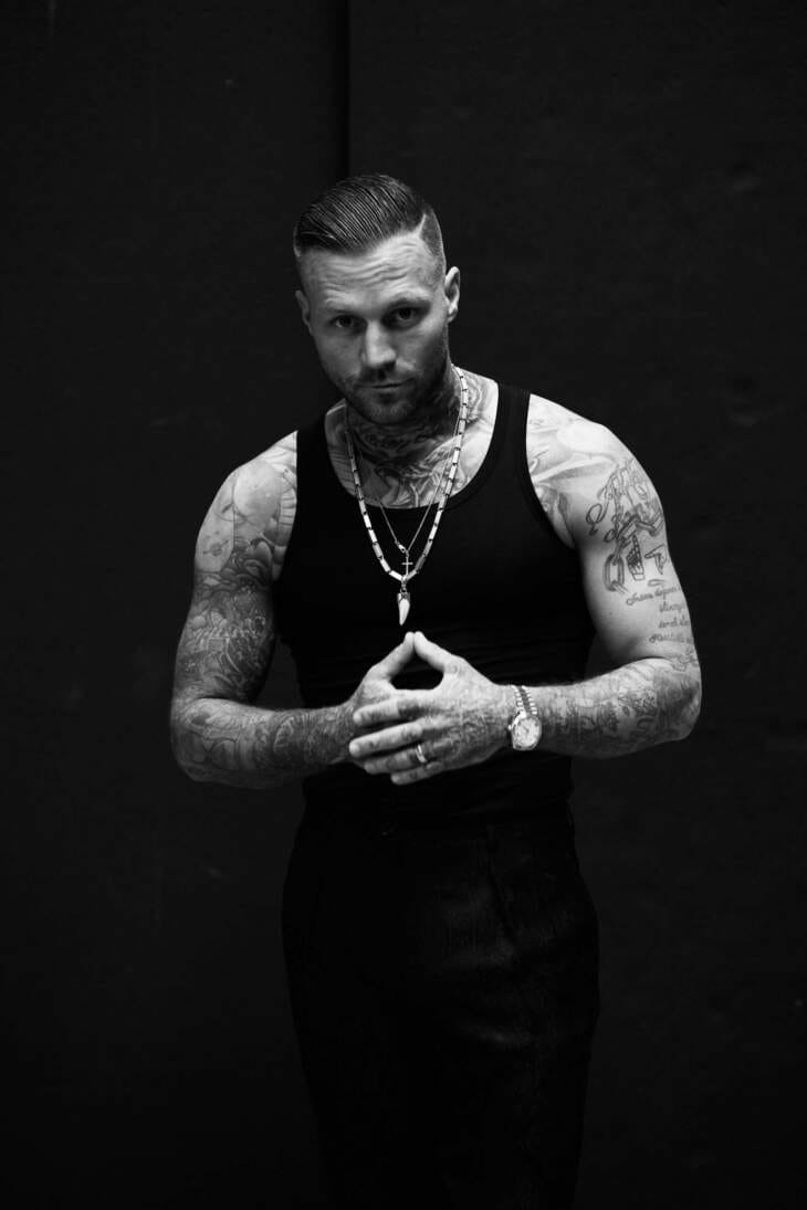 Kontra K in kraftvoller Pose mit tätowierten Armen vor dunklem Hintergrund – Berliner Rapper zeigt Entschlossenheit und Stärke, authentischer Hip-Hop-Stil, Rap-Szene Deutschland, Musik- und Streetwear-Influence Kontra K, der deutsche Rapper, posiert mit verschränkten Händen und entschlossenem Blick in einem Schwarz-Weiß-Portrait. Der Rapper trägt ein schwarzes Tanktop, seine tätowierten Arme und muskulöse Statur betonen seine Präsenz. Dieses Bild vermittelt die starke, fokussierte Persönlichkeit von Kontra K und passt perfekt zur aktuellen Tour-Ästhetik. | (c) Niculai Constantinescu