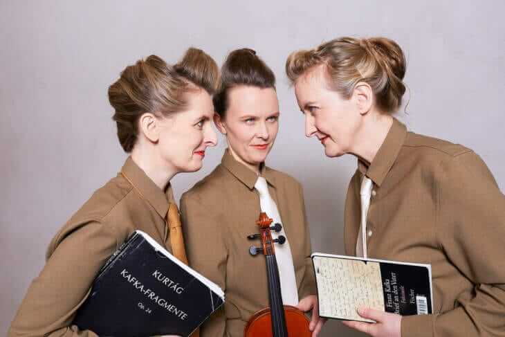 Drei Frauen in braunen Hemden halten Bücher und eine Geige, eine trägt das Buch 'Kurtág Kafka-Fragmente', eine andere 'Franz Kafka Briefe an den Vater'. Post. Kafkal Piet | Kasseler Musiktage 2024 | (c) Truhlar