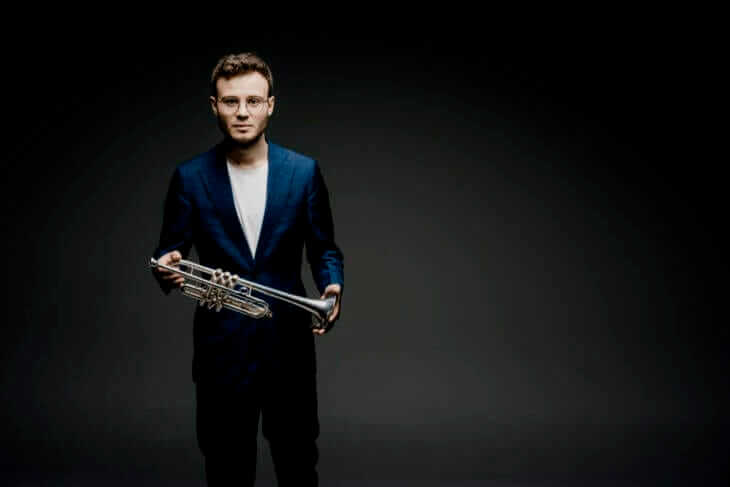 Simon Höfele, Trompeter, in dunklem Anzug mit Trompete in den Händen, vor einem dunklen Hintergrund. Simon Höfele ist einer der Gast-Stars der Kasseler Musiktage 2024 | (c) Marco Borggreve