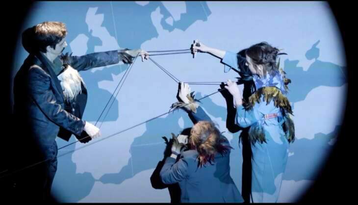 Drei Personen in Kostümen mit Federn führen eine künstlerische Performance mit Schnüren vor einer Weltkartenprojektion auf. Weltenwandern - Ein Projekt der Kasseler Musiktage 2024 | (c) Quiet City Films - Paul Holdsworth