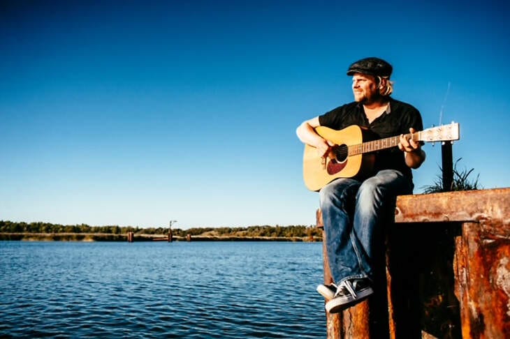 Sebel spielt Akustikgitarre und sitzt auf einem Steg am Wasser, reflektiert und nachdenklich, passend zu seinem kreativen Prozess und dem Umgang mit Emotionen. Sebel sitzt mit einer Akustikgitarre auf einem Steg am Wasser, während er in die Ferne blickt. Das Bild spiegelt die ruhige und reflektierende Seite des Musikers wider, der im Interview über seinen kreativen Prozess und den Umgang mit Emotionen spricht. | (c) Christian Thiele