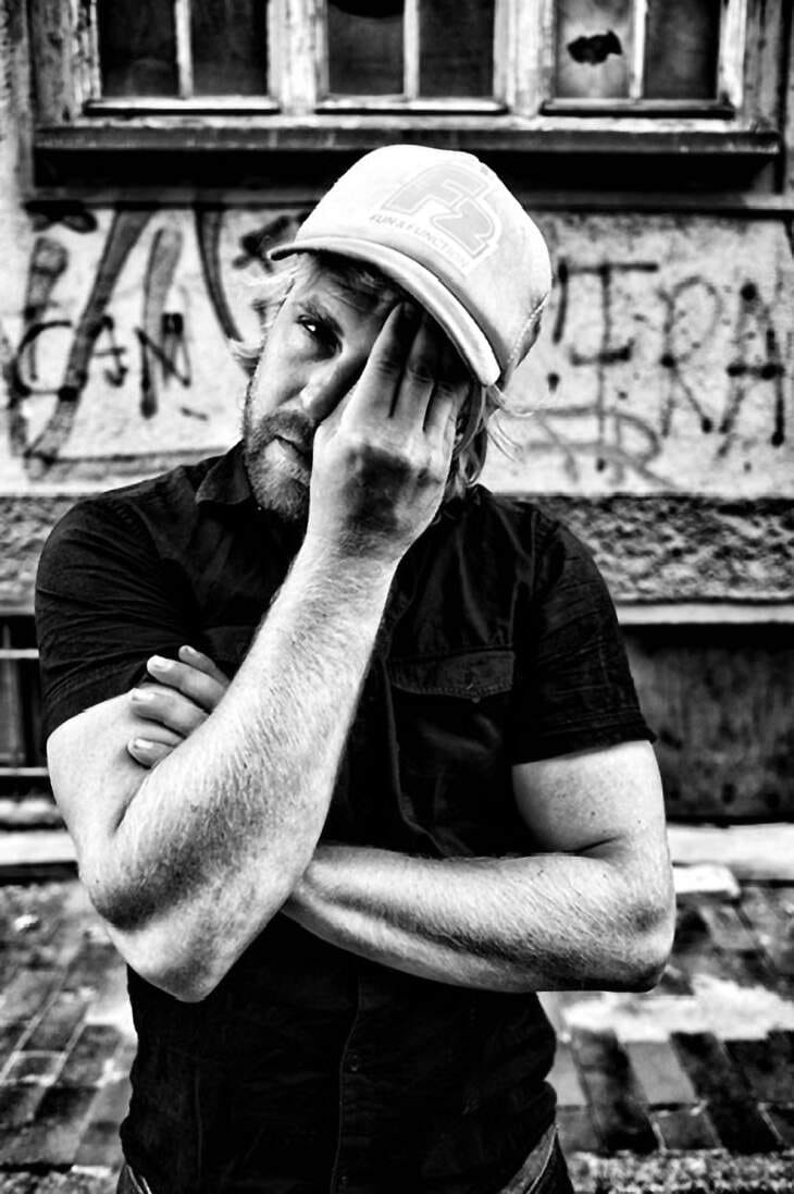 Schwarz-Weiß-Fotografie von Sebel, der nachdenklich mit einer Hand sein Gesicht berührt, während er lässig vor einer urbanen Graffiti-Wand steht. Der Musiker trägt ein T-Shirt und eine Baseballkappe, die Szene vermittelt eine rauhe, authentische Atmosphäre, passend zu seiner Persönlichkeit als Ruhrpottschnauze und Multiinstrumentalist. Sebel im Interview über sein Album 'Sträuße aus Pflaumen', Wut, Kreativität und seine musikalische Reise | (c) Christian Thiele
