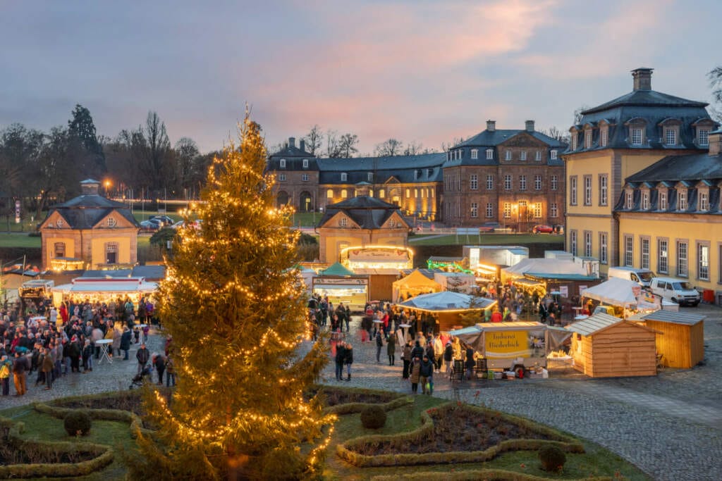 Das Bild zeigt den Weihnachtsmarkt im Innenhof eines historischen Schlosses in Bad Arolsen, aufgenommen in der Abenddämmerung. In der Mitte steht ein großer Tannenbaum, der mit warmen Lichtern geschmückt ist und festlich leuchtet. Um den Baum herum sind zahlreiche Marktstände aufgebaut, die von Menschen besucht werden. Die Stände sind mit Lichtern dekoriert und bieten verschiedene Waren an. Im Hintergrund erheben sich die gelb beleuchteten Gebäude des Schlosses, die den Weihnachtsmarkt umrahmen und eine gemütliche, festliche Atmosphäre schaffen. Die Szene vermittelt eine vorweihnachtliche Stimmung in einer idyllischen Umgebung. Weihnachtsmarkt Bad Arolsen 2024: Das Bild zeigt den Weihnachtsmarkt im Innenhof eines historischen Schlosses in Bad Arolsen, aufgenommen in der Abenddämmerung. In der Mitte steht ein großer Tannenbaum, der mit warmen Lichtern geschmückt ist und festlich leuchtet. | (c) HB&B