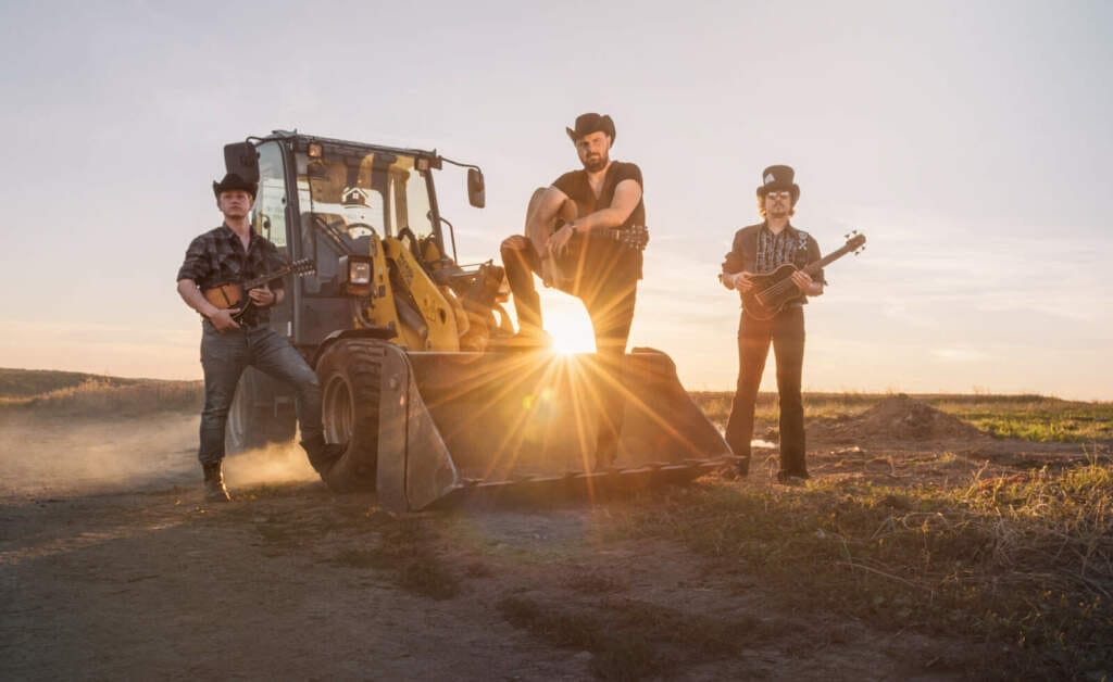 Country-Band-Posing vor einem Bagger bei Sonnenuntergang auf einer staubigen Landstraße – rustikale Stimmung mit Mandoline und Gitarre Mandowar sind startbereit für ihr Konzert zum 30. Jubiläum der Freien Musikinitiative Schrecksbach | (c) Stephan Klement