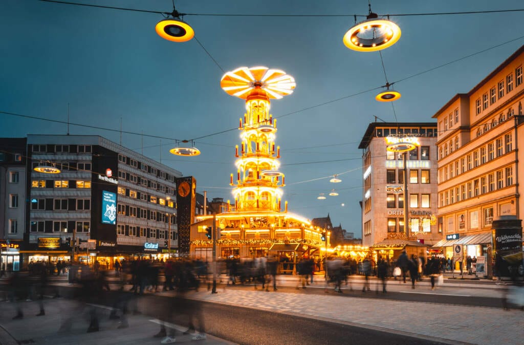 Beleuchtete Weihnachtspyramide auf dem Weihnachtsmarkt in Bielefeld bei Dämmerung 2023 hat der Bielefelder Weihnachtsmarkt ein neues Highlight bekommen: die 22 Meter hohe Weihnachtspyramide auf dem Jahnplatz. Sie zählt zu den größten Pyramiden in ganz Deutschland und ist die größte in Nordrhein-Westfalen. | (c) Tim-Frohlich