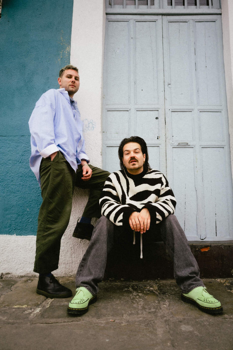 Zwei Männer in Streetwear vor blauer Wand und Holztür, Milky Chance posieren vor blauer Tür, modern gekleidet. Milky Chance und das Kasseler Staatsorchester werden in 2025 in der Karlsaue in Kassel: Ein musikalisches Highlight des Stadtsommers 2025 | Anthony Molina