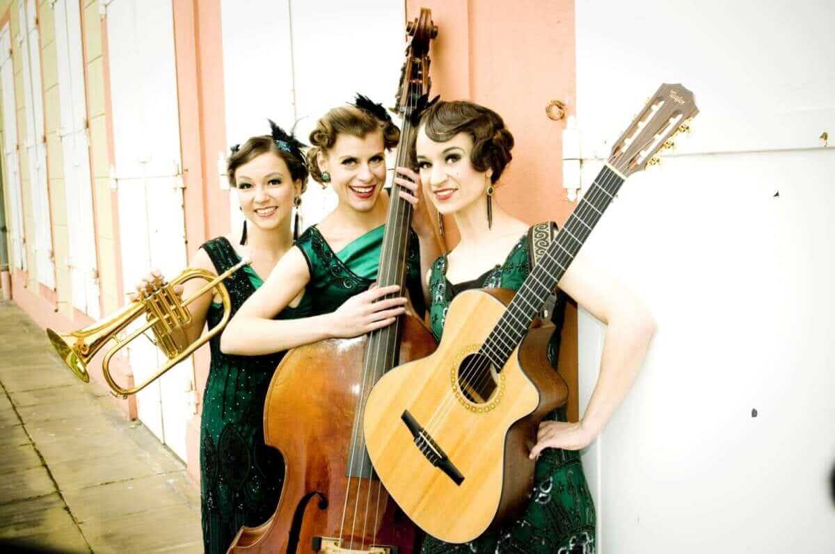 Swing, Charme und ein Hauch Nostalgie: Die Zucchini Sistaz posieren gut gelaunt mit Trompete, Kontrabass und Gitarre vor heller Kulisse. In abgestimmten Vintage-Kleidern verkörpern sie musikalische Leichtigkeit – ein stimmiger Vorgeschmack auf ihren Auftritt beim „Sommer im Park 2026“ in Vellmar. Zucchini Sistaz | (c) Katharina Tenberge