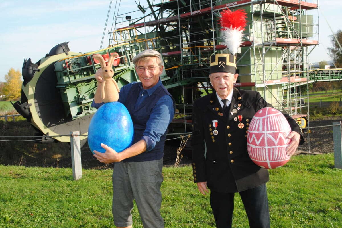 Zwei Männer mit Ostereiern stehen vor einem Schaufelradbagger des Bergbaumuseums Frühlingserwachen im Themenpark Kohle & Energie des Hessischen Braunkohle Bergbaumuseums in Borken | (c) Stadt Borken