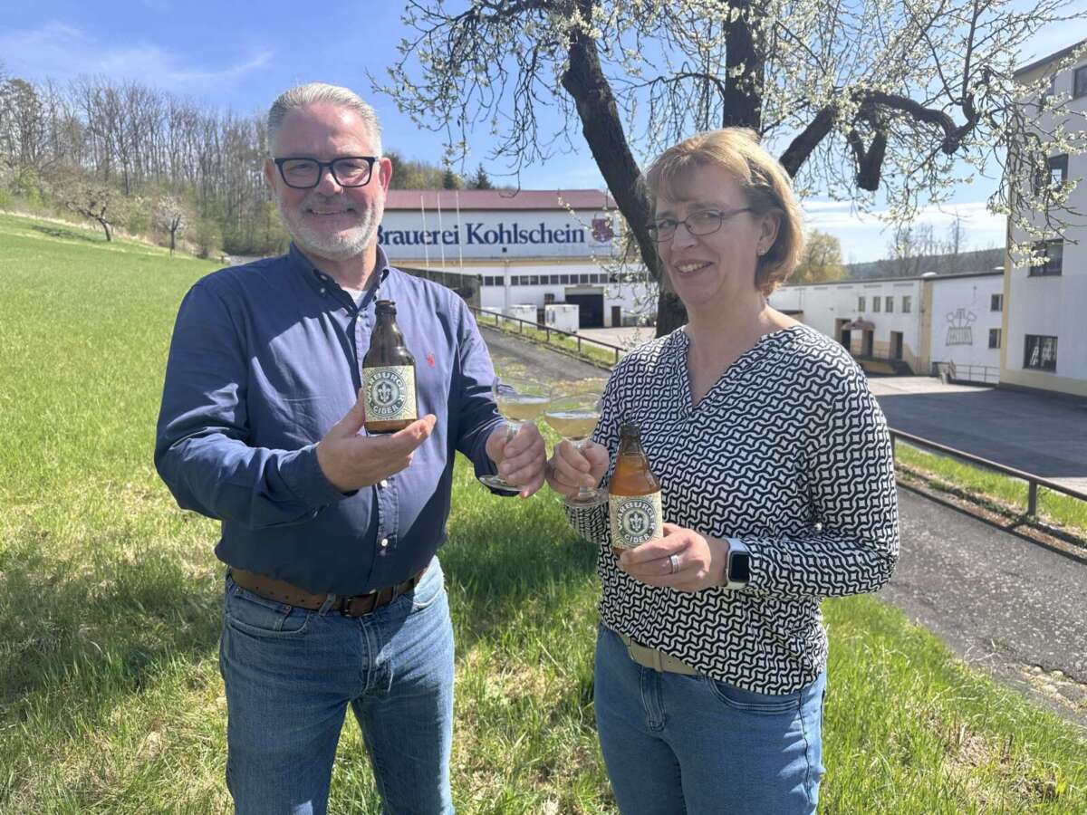 Zwei Personen stoßen mit Apfel-Cider vor Brauerei Kohlschein an Die Warburger Brauerei Kohlschein bringt 2025 ihren beliebten „White-Cider“ zurück in den Handel – frisch, spritzig und perfekt für den Frühling: Bei Sonnenschein wird der neue Jahrgang direkt vor Ort verkostet. | (c) Warburger Brauerei