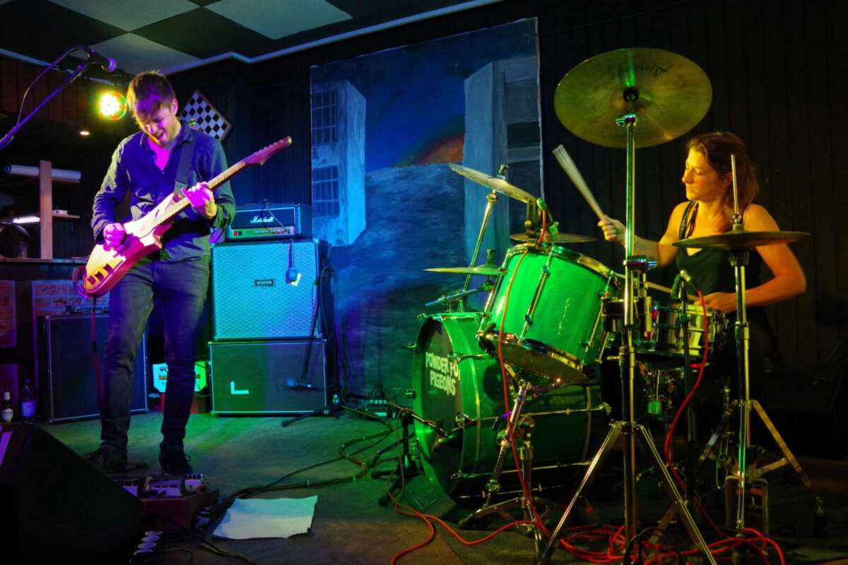 Links im Bild ist Rhys von Powder For Pigeons zu sehen, wie er Gitarre spielt. Rechts sitzt Maike am Schlagzeug. Die Bühne ist mit grünem und blauem Licht ausgeleuchtet. Powder For Pigeons rockten bereits zuvor die Bühne in Marsberg | Live im JBZ am 27.09.2019 | (c) JBZ