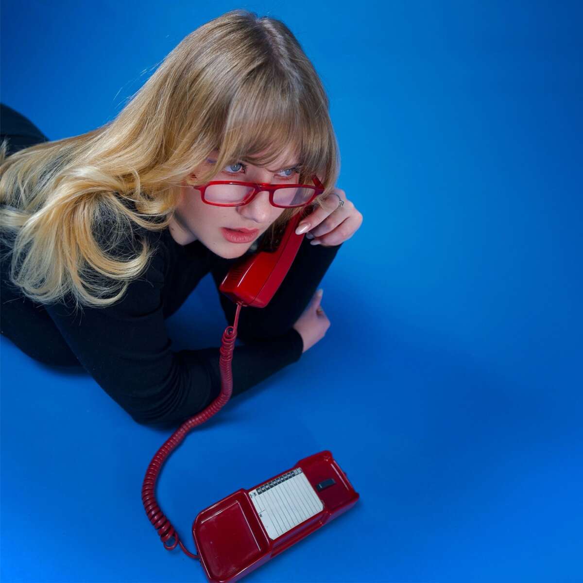 EIne blonde Frau mit roter Brille spricht in ein rotes Telefon. Der Hintergrund ist blau. Dilla ist am Telefon – sie will wissen ob du dieses Jahr auch beim Sommerfestival dabei bist | (c) Lola Schreiner