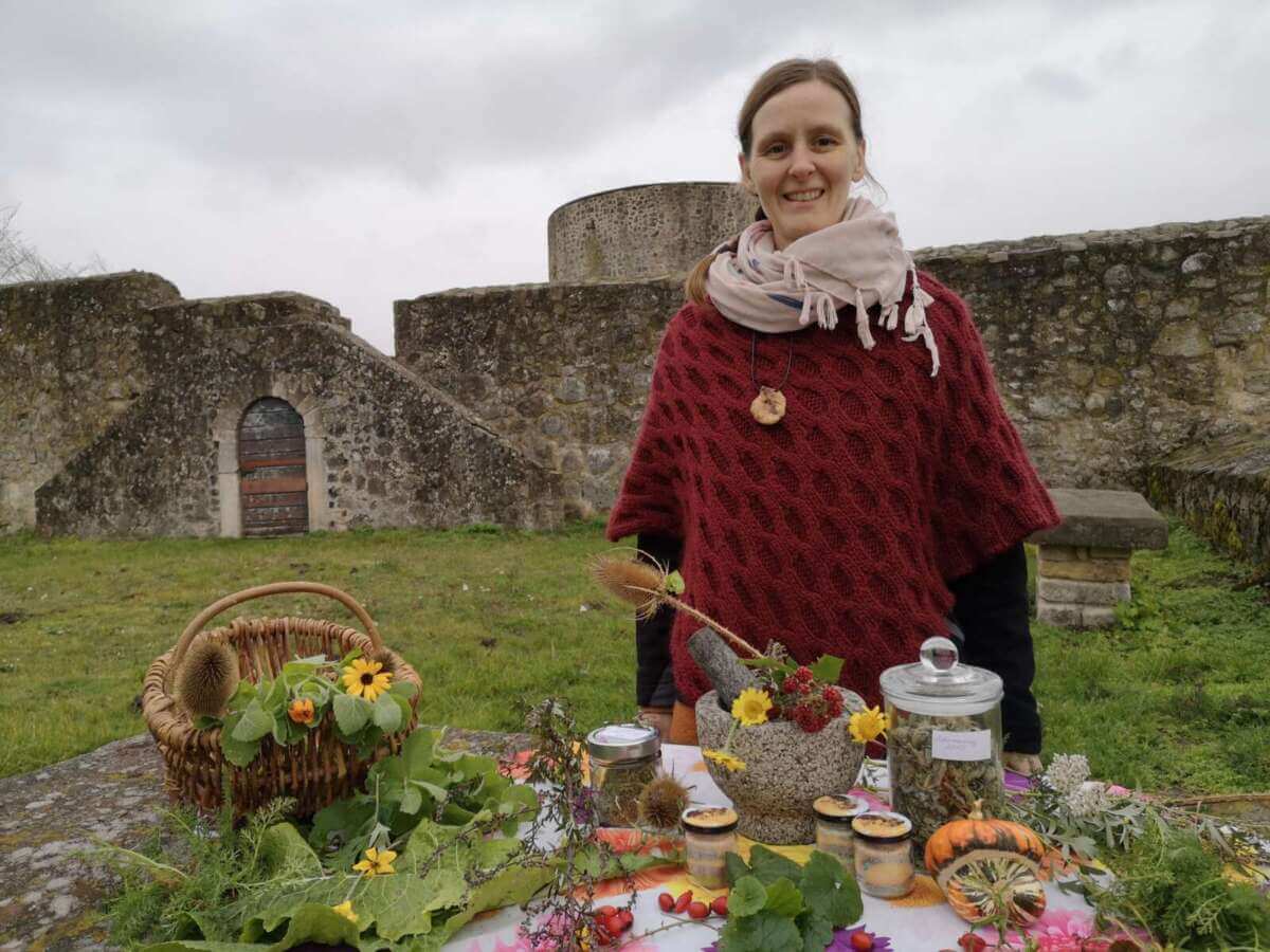 Bei einer Kräuterwanderung an Hessens Krone präsentiert Anke Dobel einen Tisch voller Pflanzen, Körbe und Mörser. Sie lächelt in die Kamera, während im herbstlichen Hintergrund eine Burg zu sehen ist. Bei herbstlicher Kulisse an Hessens Krone zeigt Kräuterfrau Anke Dobel einen Tisch voller Pflanzen, Körbe und Mörser. Mit einem Lächeln lädt sie dazu ein, die Welt der Kräuter durch Ihre Kräuterwanderung zu entdecken | (c) Tourismusgesellschaft Melsunger Land mbH