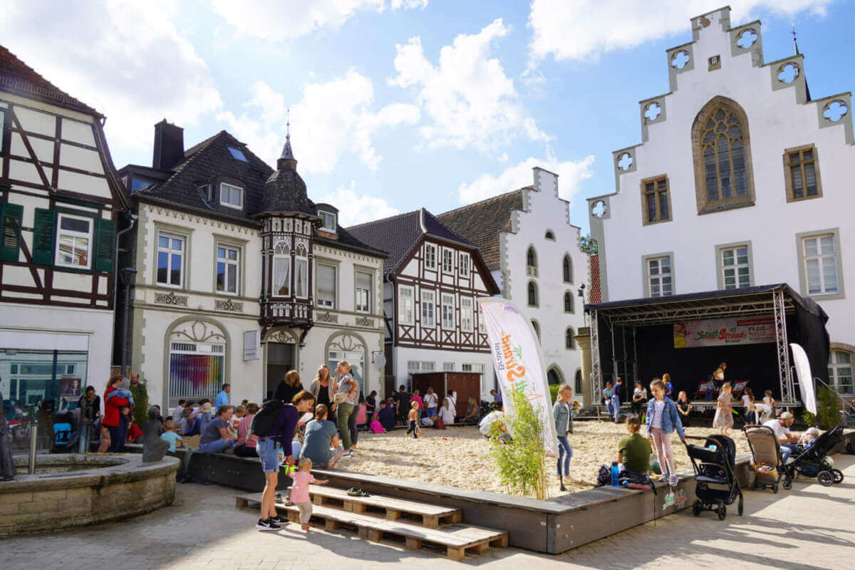 In der Brakeler Innenstadt reihen sich charmante Fachwerkhäuser aneinander, während am Stadtstrand unter sonnigem Himmel und im Schatten des historischen Rathauses entspanntes Leben pulsiert. Stadtstrand Brakel 21.08.2024 | © Kita Hembsen