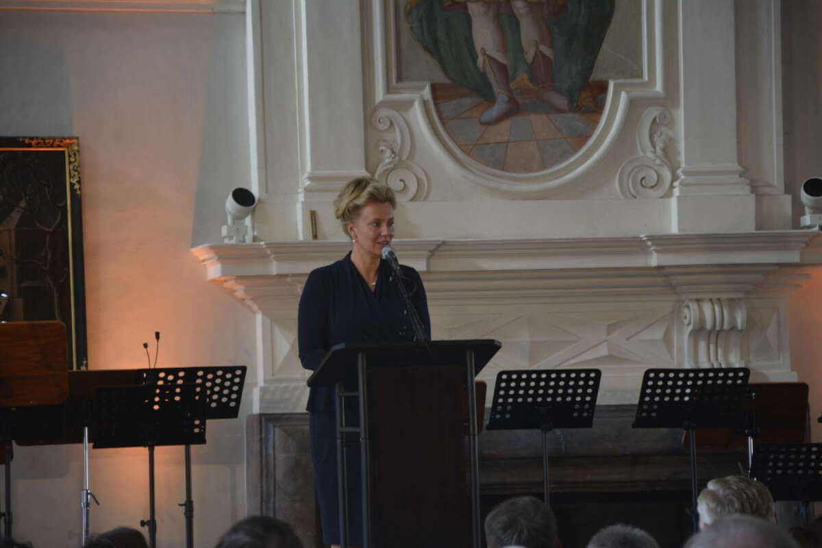 Grußwort von Ina Brandes, Ministerin für Kultur und Wissenschaft des Landes NRW, beim Via Nova Kunstfest 2025 im Schloss Corvey (UNESCO-Weltkulturerbe, Höxter). Auf der Bühne steht ein Pult, der prunkvolle Saal ist mit Stuck und Gemälden verziert, das Publikum sitzt aufmerksam und verfolgt die Rede.