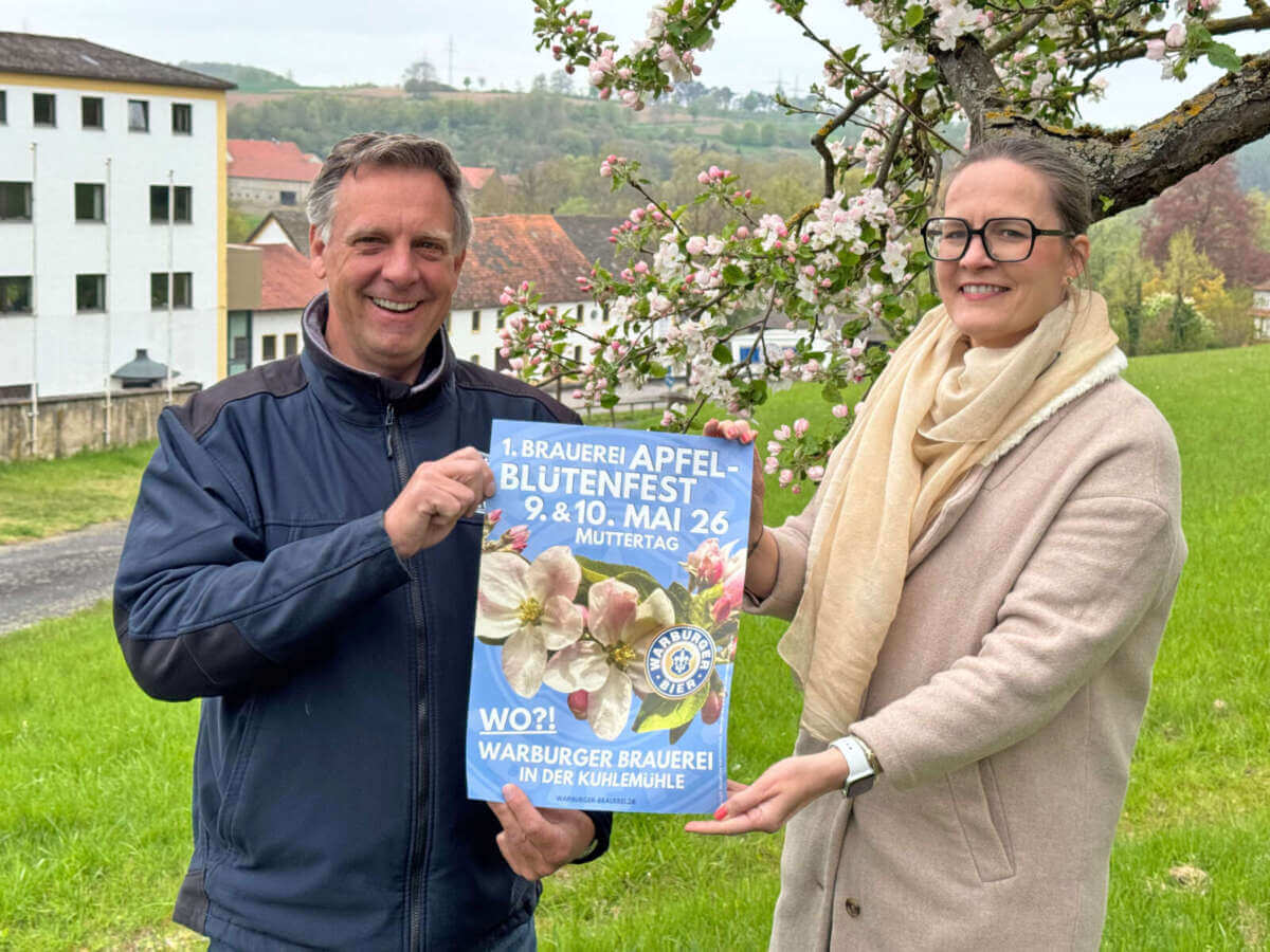 Franz-Axel Kohlschein (Geschäftsführer) und Valeria Geritzen (Marketing) präsentieren auf einer Wiese vor blühendem Apfelbaum vor der Brauerei ein Plakat für das 1. Brauerei Apfelblütenfest am 9. und 10. Mai 2026 in Warburg. | (c) Wildwechsel.de Brauerei Apfelblütenfest in Warburg 2026 | (c) Wildwechsel.de