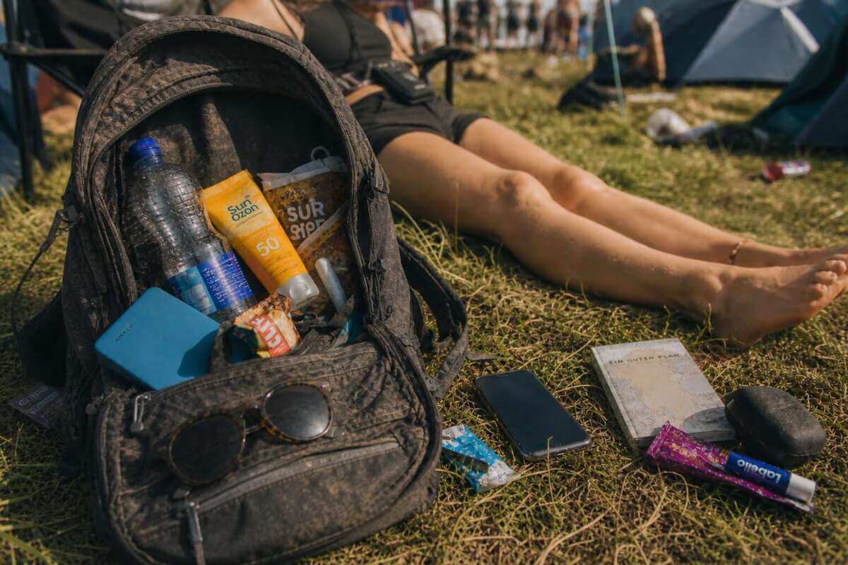 Ein geöffneter Rucksack liegt auf einer Wiese auf einem Festival-Campingplatz. Darin befinden sich eine Wasserflasche, Sonnencreme, eine Powerbank, Snacks und weitere persönliche Gegenstände. Neben dem Rucksack liegen ein Smartphone und kleine Alltagsartikel im Gras. Im Hintergrund sind unscharf Zelte und Menschen zu sehen, eine Person liegt entspannt auf dem Boden. Festival Essentials im geöffneten Rucksack mit Wasserflasche, Sonnencreme, Powerbank, Snacks und Smartphone auf dem Campingplatz