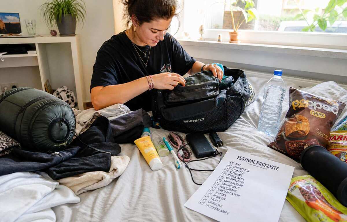 Eine Person sitzt auf einem Bett und packt einen Rucksack für ein Festival. Um sie herum liegen verschiedene Gegenstände wie Kleidung, ein Schlafsack, eine Wasserflasche, Snacks, eine Powerbank und Hygieneartikel. Auf dem Bett liegt außerdem eine ausgedruckte Festival-Packliste mit abgehakten Punkten. Tageslicht fällt durch ein Fenster in den Raum. Festival Tasche packen zuhause mit Packliste, Kleidung, Schlafsack, Wasserflasche, Snacks und Powerbank