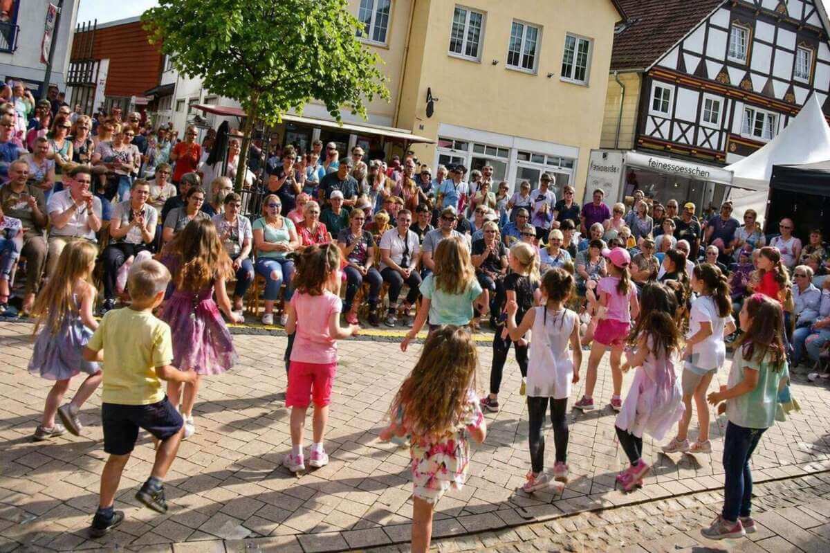 Fröhliche Kinder tanzen und spielen vermutlich auch beim Stadtfest Brakel 2026, während zahlreiche Zuschauer das bunte Programm verfolgen – ein lebendiger Treffpunkt für Familien mitten in der Brakeler Innenstadt. | Ki-generiert - (c) Wildwechsel.de
