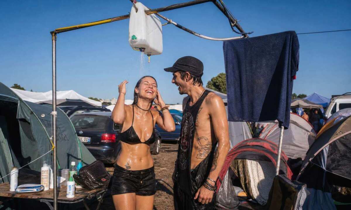 Zwei junge Erwachsene stehen auf einem Festival-Campingplatz unter einer improvisierten Dusche, die aus einem Wasserkanister oder einer mobilen Campingdusche besteht. Sie tragen teilweise noch Kleidung wie Shorts oder ein T-Shirt. Neben ihnen liegen ein Handtuch und Hygieneartikel wie Shampoo oder Duschgel. Im Hintergrund sind Zelte und weitere Festivalbesucher zu sehen. Die Umgebung wirkt staubig und sommerlich. Festival Dusche auf dem Campingplatz mit improvisierter Campingdusche, Handtuch und Hygieneartikel