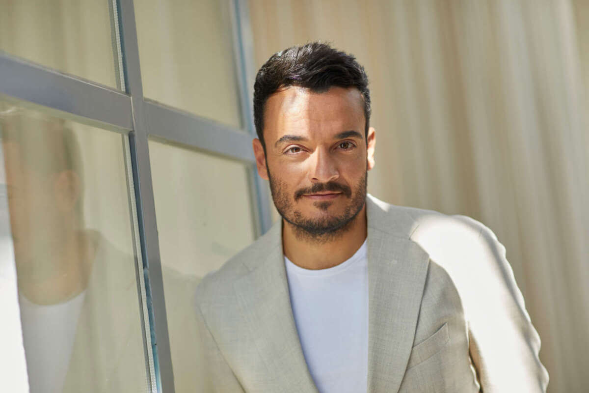 Das Bild zeigt Giovanni Zarrella in einem hellen Sakko und weißem Shirt. Er steht an einem Fenster, sein Gesicht ist weich beleuchtet, links spiegelt sich seine Silhouette. Giovanni Zarrella ist einer der Künstler beim Butzbach Open Air 2026 und blickt in hellem Sakko in die Kamera. | (c) Boris Breuer