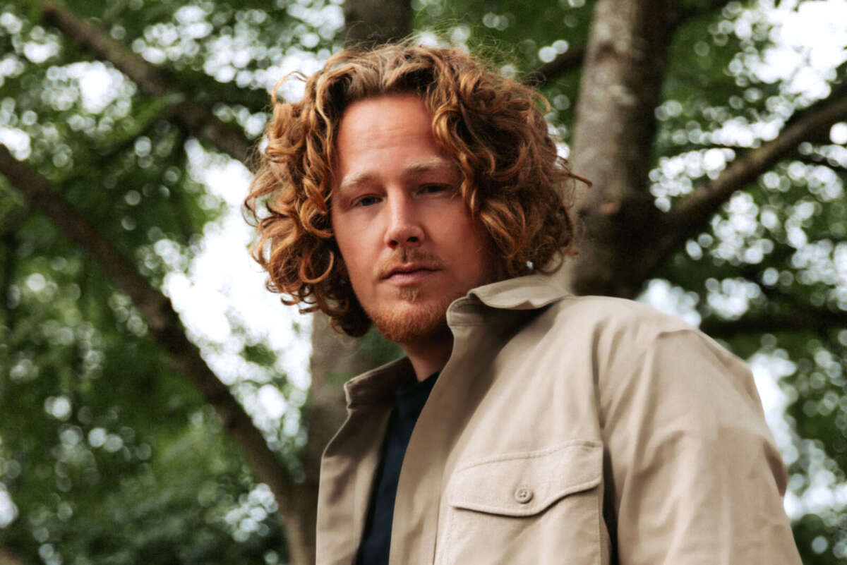 Michael Schulte steht im Freien vor dichtem Grün und Baumstämmen. Er trägt eine helle Jacke, schaut ruhig in die Kamera und wirkt passend zum Pop-Fokus des Lippe Open Air 2026 nahbar und konzentriert. Porträt von Michael Schulte vor Bäumen, angekündigt für das Lippe Open Air 2026
