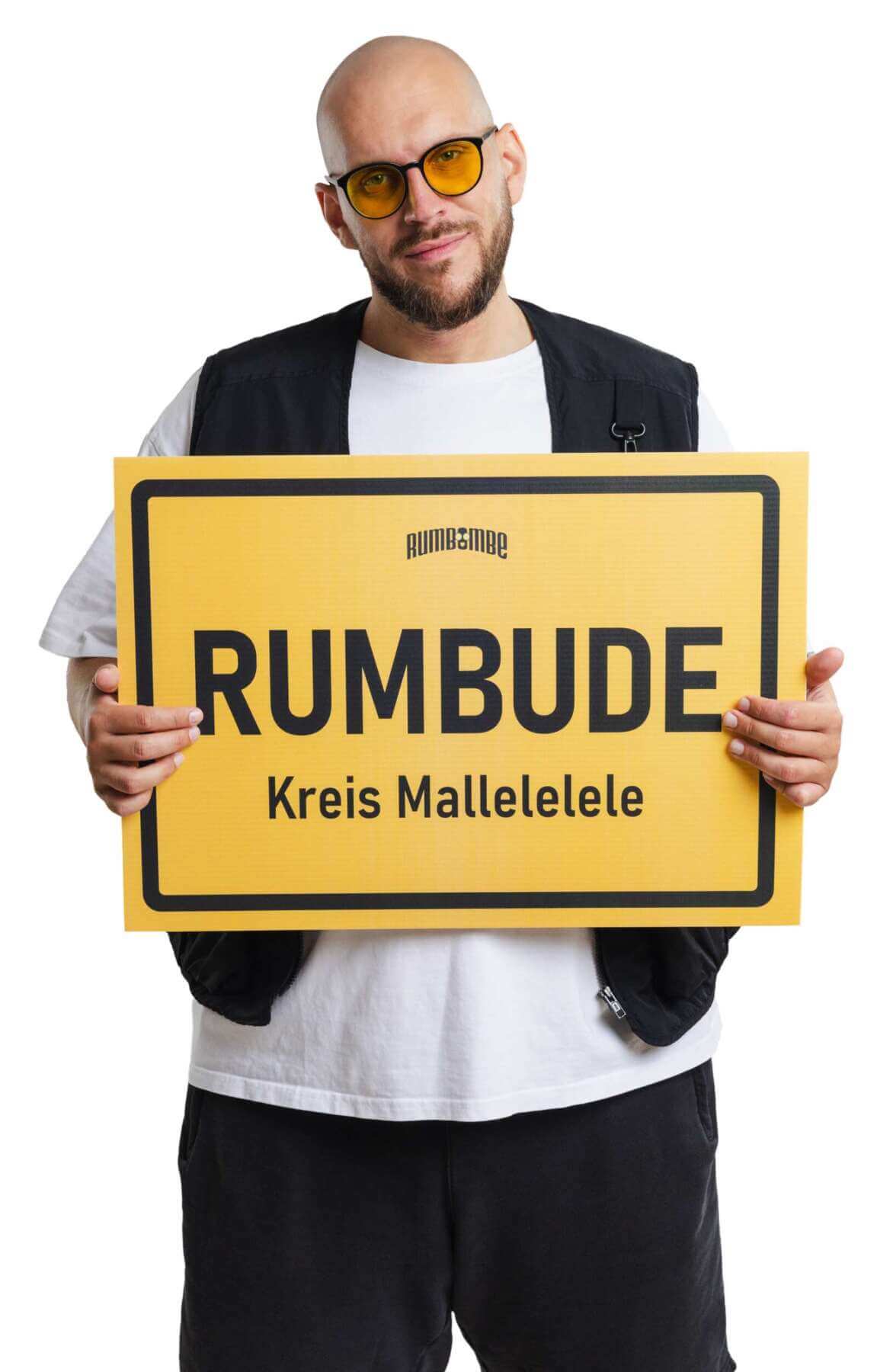 Rumbombe hält ein gelbes Ortsschild mit der Aufschrift „Rumbude“ vor hellem Hintergrund. Der Partyschlager-Künstler tritt bei der Warburger Oktoberwoche 2026 auf. (c) Markus Krampe Booking