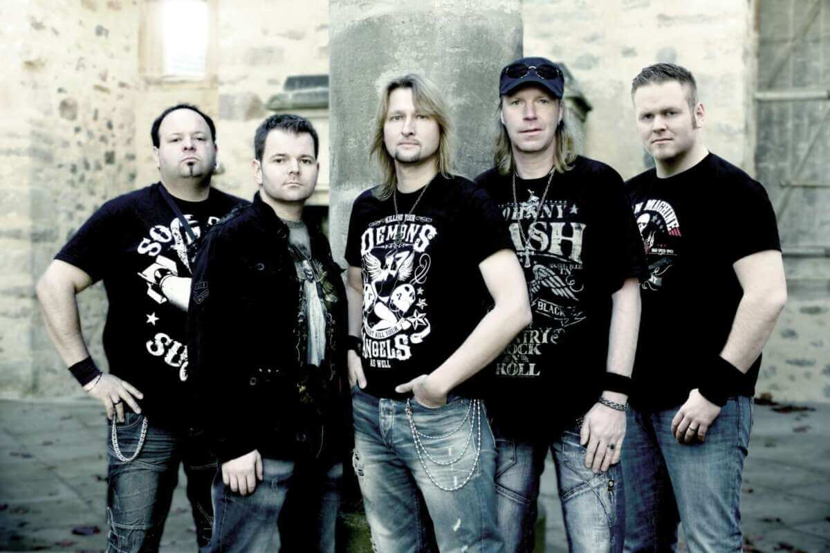 Fünf Mitglieder der Band Wild Night stehen nebeneinander vor einer historischen Steinmauer und einem Säulenelement. Die Musiker tragen überwiegend schwarze Kleidung mit Band- und Rockmotiven. Einige haben die Hände in den Taschen oder verschränken die Arme. Die Aufnahme ist als Gruppenporträt gestaltet und zeigt die Band, die im Rahmen der Ziegenhainer Salatkirmes auftritt. Gruppenfoto der Band Wild Night, die bei der Ziegenhainer Salatkirmes in Schwalmstadt auftritt.