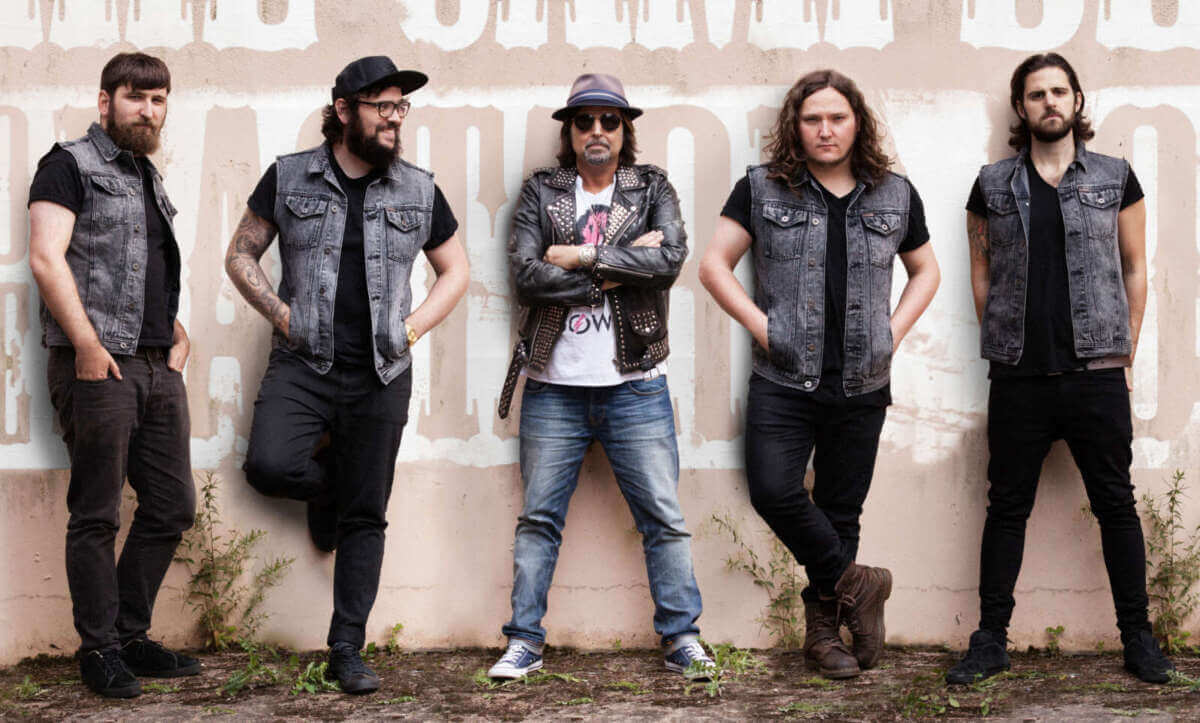 Fünf Musiker stehen nebeneinander vor einer hellen Wand mit großflächigen Schriftzügen. In der Mitte steht Phil Campbell mit Hut, Sonnenbrille, Lederjacke und verschränkten Armen. Die vier weiteren Bandmitglieder tragen überwiegend schwarze Kleidung und Jeanswesten, einige haben lange Haare oder Bärte. Mehrere lehnen entspannt an der Wand. Das Bild vermittelt eine lockere, klassische Rockband-Atmosphäre. Bandfoto von Phil Campbell and the Bastard Sons vor einer Mauer. Die Hardrock-Band tritt beim Rock N Revel 2026 auf. | (c) Phil Campbell