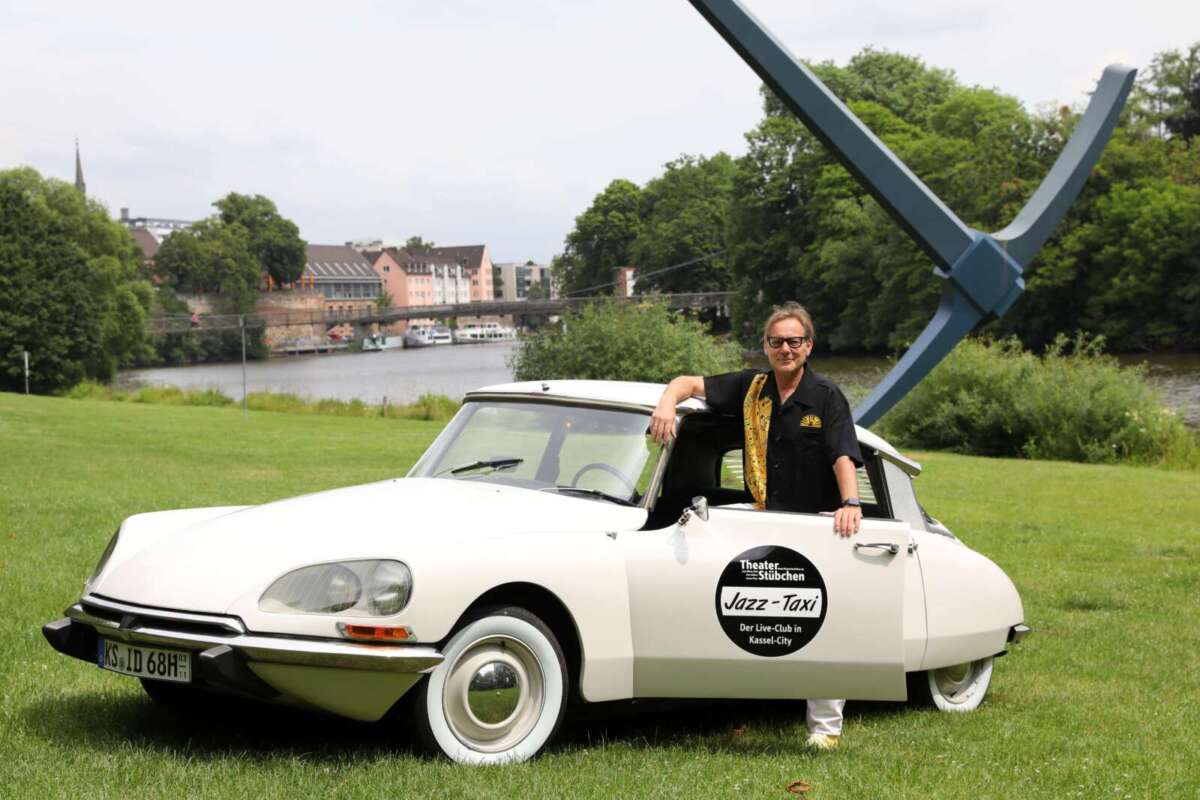 Auf einer grünen Wiese steht ein weißer klassischer Citroën mit geöffneter Fahrertür. Ein Mann lehnt am Fahrzeug. Auf der Tür befindet sich ein schwarzer runder Schriftzug mit der Aufschrift „Jazz-Taxi – Der Live-Club in Kassel-City“. Im Hintergrund sind Wasser, Bäume, Häuser und eine große abstrakte Metallskulptur zu sehen. Historischer weißer Citroën als Jazz-Taxi des Theaterstübchens vor Kasseler Fluss- und Parkkulisse.