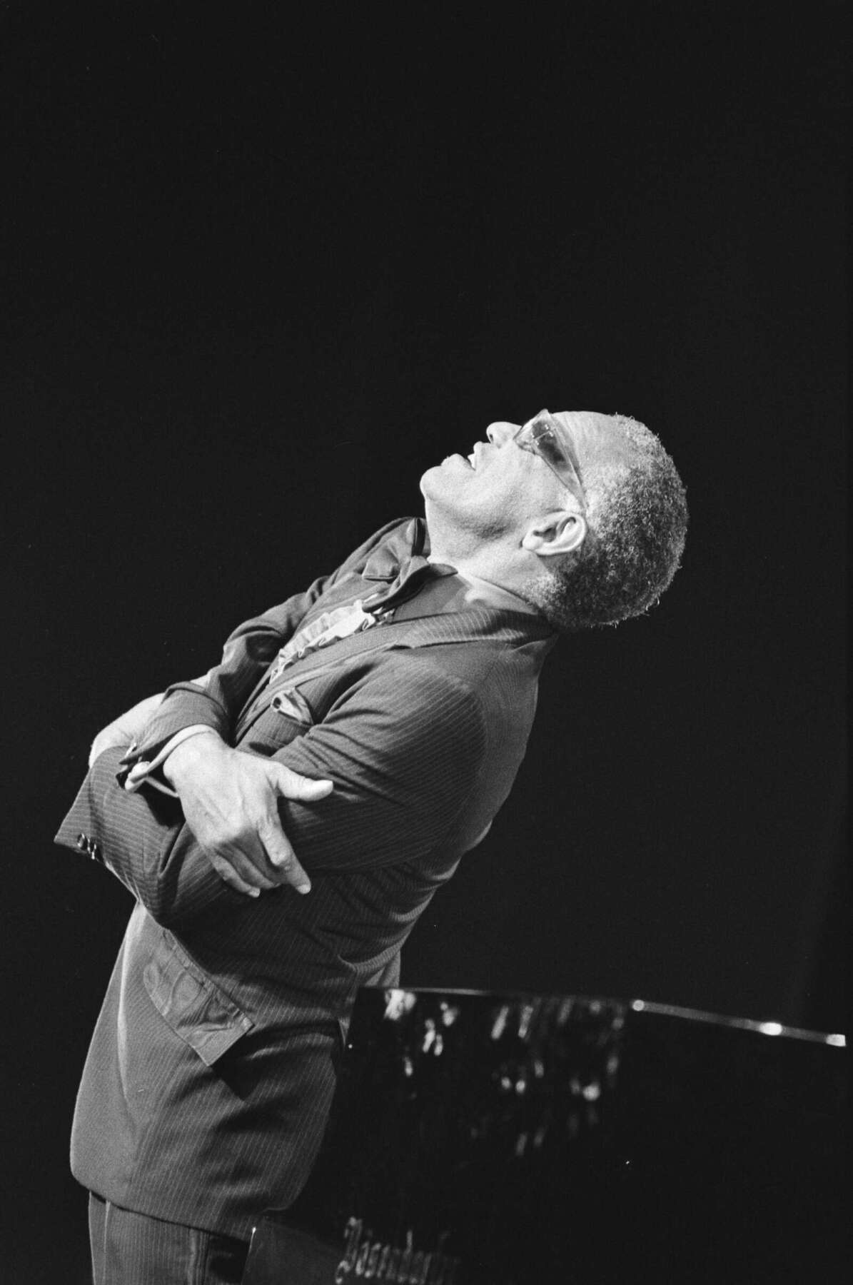 Die Schwarzweißaufnahme zeigt Ray Charles auf einer dunkel gehaltenen Bühne. Er trägt einen Anzug und eine Sonnenbrille, hat die Arme vor der Brust verschränkt und lehnt seinen Oberkörper deutlich nach hinten. Sein Kopf ist weit zurückgelegt und nach oben gerichtet. Rechts unten ist ein Teil des glänzenden Flügels zu sehen. Der fast vollständig schwarze Hintergrund hebt den Musiker und seine ausdrucksstarke Körperhaltung stark hervor. Ray Charles lehnt sich bei einem Auftritt am Flügel weit zurück, die Arme vor der Brust verschränkt – Schwarzweißaufnahme vom North Sea Jazz Festival 1983.