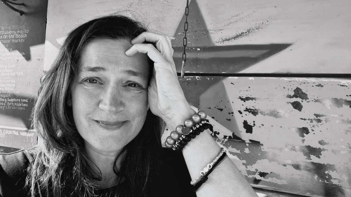 Schwarz-Weiß-Porträt von Ruth Hebler. Sie sitzt vor einer Wand mit großem Sternmotiv und blickt in die Kamera. Ihr langes dunkles Haar fällt offen über die Schultern. Eine Hand liegt an ihrer Stirn; am Handgelenk trägt sie mehrere Armbänder. Schwarz-Weiß-Porträt der Cartoonistin Ruth Hebler zur Ausstellung „Alles wird Ruth“ in Kassel
