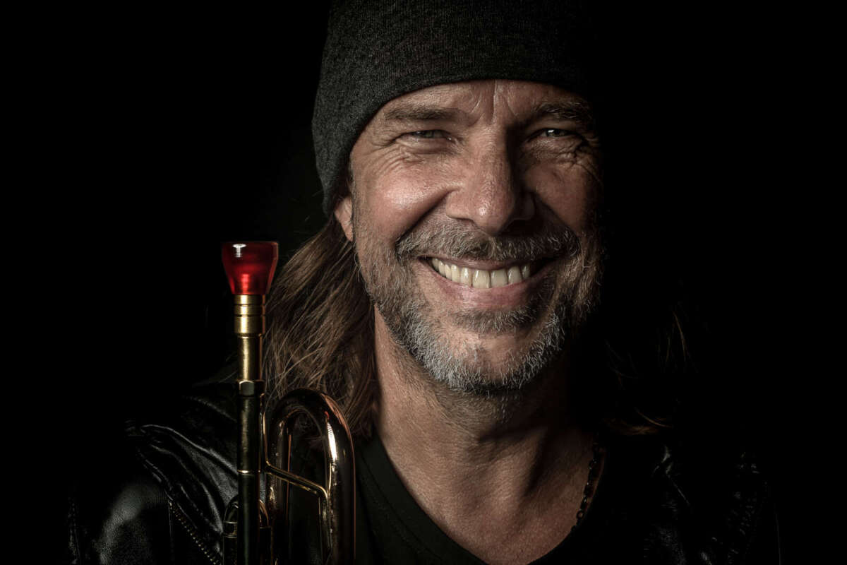 Rüdiger Baldauf mit Trompete vor schwarzem Hintergrund. Das Bild gehört zum Wildwechsel-Artikel über Rüdiger Baldauf feat. Max Mutzke und die Trumpet Night im Anthroposophischen Zentrum Kassel. Foto: Simon Engelbert. Porträt von Rüdiger Baldauf mit schwarzer Mütze und Trompete auf einem Pressefoto zur Trumpet Night